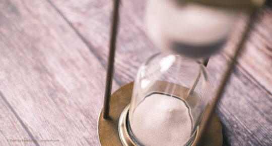 Eine Sanduhr steht auf Holzbalken. Der weiße Sand rinnt hinunter. © Towfiqu barbhuiya – unsplash.com