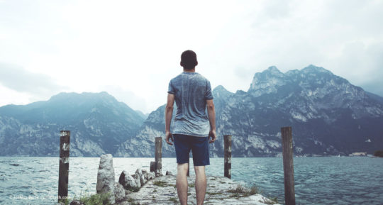 Mann steht auf einem Steg an einem See und schaut auf Berge © Joshua Earle – unsplash.com