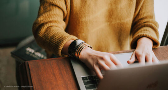 Frau in gelbem Pullover sitzt am Notebook und tippt. © Christin Hume – unsplash.com